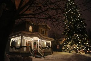 a huge spruce tree lit up with white lights under a recent snowfall. The decoration of green wreaths and swags on the house with the Christmas tree. This is a great example when you prepare your home exterior for the holidays